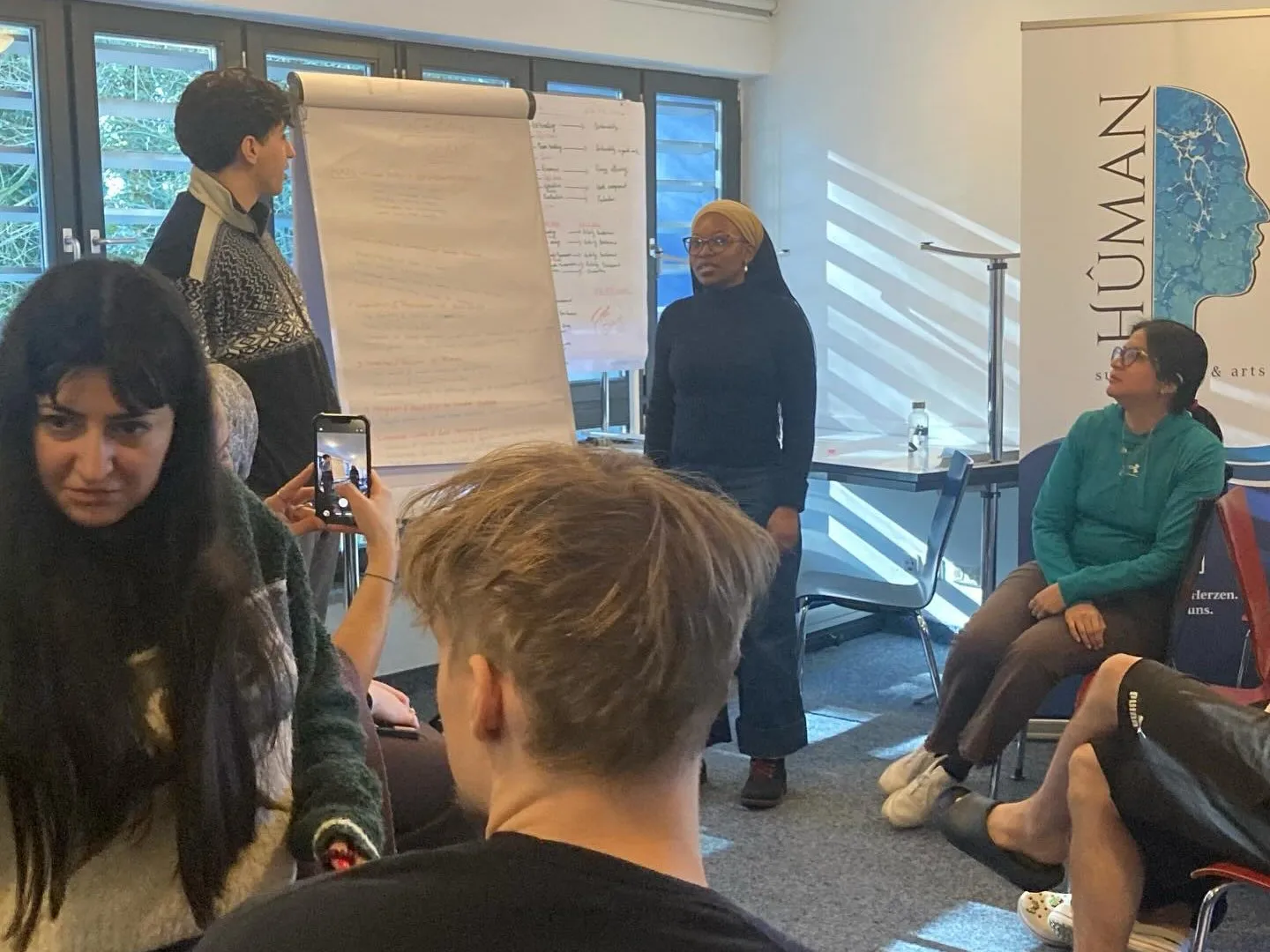 Eine Teilnehmerin von Sustain Youth stellt Ergebnisse am Flipchart vor, die Gruppe hört zu; rechts der Hûman-Aufsteller
